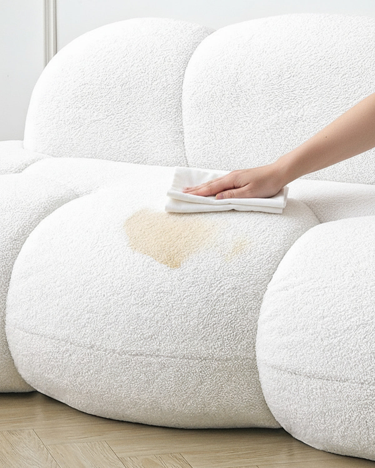 Une main tamponne une tache légère sur l'assise d'un canapé nuage en tissu bouclé, avec un chiffon de coton blanc